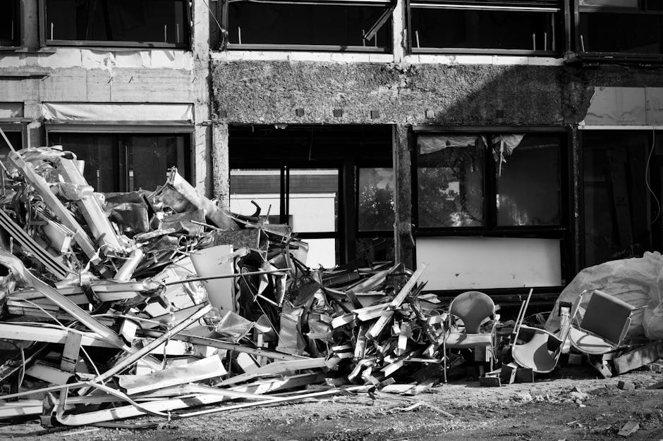 A large pile of metal debris and broken furniture, including chairs and fragments of construction materials, is situated in front of a partially damaged building with broken windows and exposed concrete walls. The debris appears to consist of twisted metal, shattered roofing panels, and assorted discarded objects, creating a chaotic scene on a paved surface outside a deteriorating commercial or institutional structure. The environment suggests a recent demolition or on-site clearance, with some items leaning or stacked haphazardly amid the wreckage. Behind the pile, the building's windows are either shattered or missing, revealing dark interior spaces and construction scaffolding or external framing. The natural lighting is neutral, with shadows cast by the structure and debris, emphasizing the stark contrast between the debris and the surrounding environment. This scene aligns with independent rubbish removal or waste handling processes, akin to those provided by companies such as Waste Collection Uxbridge, specializing in clearing bulky debris and construction waste from such sites.