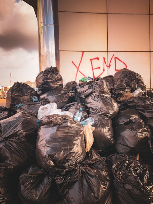 A large pile of black rubbish bags filled with waste materials is stacked against a tiled wall in an outdoor urban setting. Some bags are partially torn or bulging, revealing glimpses of their contents, including plastic bottles and packaging. The bags are clustered together on a paved surface, with a few loose items like plastic bottles and small debris scattered around the base. The wall behind the pile features a bold red graffiti message reading 'X END' painted directly on the tiles, which are light-colored with visible grout lines. The scene is illuminated by natural daylight, with a partly cloudy sky visible in the top left corner, suggesting a city environment. This image depicts a typical example of private rubbish collection and on-site waste clearance, similar to services provided by waste collection specialists for non-regular waste removal needs.