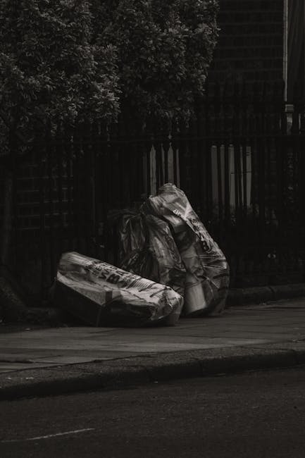A large pile of metal debris and broken furniture, including chairs and fragments of construction materials, is situated in front of a partially damaged building with broken windows and exposed concrete walls. The debris appears to consist of twisted metal, shattered roofing panels, and assorted discarded objects, creating a chaotic scene on a paved surface outside a deteriorating commercial or institutional structure. The environment suggests a recent demolition or on-site clearance, with some items leaning or stacked haphazardly amid the wreckage. Behind the pile, the building's windows are either shattered or missing, revealing dark interior spaces and construction scaffolding or external framing. The natural lighting is neutral, with shadows cast by the structure and debris, emphasizing the stark contrast between the debris and the surrounding environment. This scene aligns with independent rubbish removal or waste handling processes, akin to those provided by companies such as Waste Collection Uxbridge, specializing in clearing bulky debris and construction waste from such sites.
