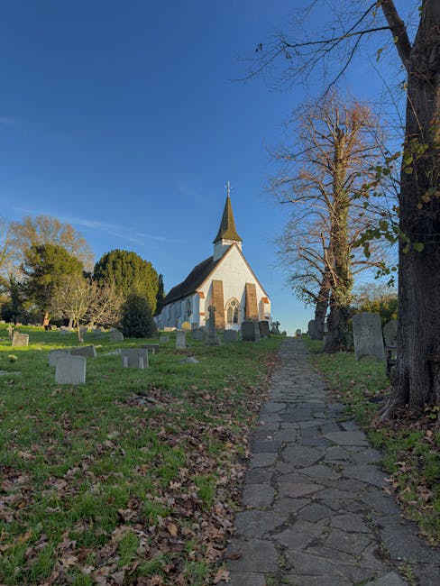 A narrow, irregular stone pathway leading up a gentle incline through a grassy area scattered with fallen leaves, flanked by old, leafless trees on the right and a few smaller trees and bushes on the left. In the background, there is a small white church with a pointed steeple topped by a weather vane, situated among several mature trees and old gravestones arranged around the churchyard, indicative of a rural or historical setting. The sky above is clear and bright blue with minimal clouds, suggesting a calm, sunny day. The scene is captured during daytime, with natural sunlight casting soft shadows, and the overall atmosphere reflects a peaceful, open countryside environment suitable for private outdoor visits or local heritage preservation efforts, aligning with independent waste collection and clearance services in the area.