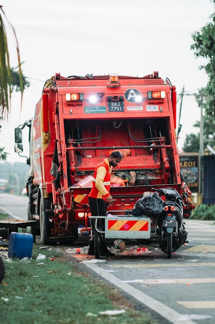 A worker dressed in a red and yellow uniform is operating a large red refuse collection truck on the side of a street, engaging in rubbish removal. The back of the truck is open, revealing mechanical components and a loading area, with some debris visible on the ground beneath. The worker appears to be handling waste or adjusting equipment near a black bag placed on a small platform attached to the truck. In the background, blurred trees and utility poles line the street, which is paved and marked with white lines, indicating a designated parking or loading zone. Nearby, a small motorbike is parked close to the waste collection vehicle, and there are various items such as a blue container and other scattered debris nearby, suggesting an on-site clearance operation. The scene is illuminated by natural daylight, with overcast skies providing diffuse lighting, and the environment reflects typical urban waste collection activity, which could be part of an independent rubbish disposal or private waste management service related to rubbish removal in the local area.